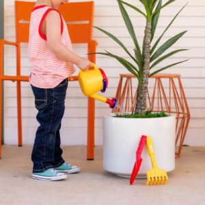 Child watering a potted plant with a Spielstabil yellow watering can. Next to the potted plant on the ground are a red plastic shovel and a yellow plastic rake.