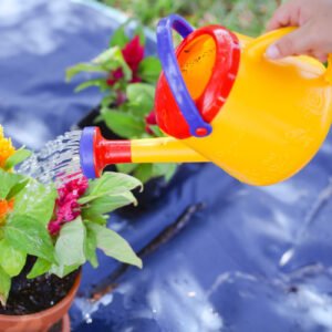 A hand holding a colorful Spielstabil watering can pouring water on plants with green leaves and vibrant flowers.