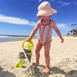 Child playing on a sunny beach with a sand funnel toy and shovel.
