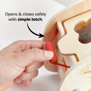 Close-up of a hand opening a simple red latch on a wooden toy shape sorter. Text reads 